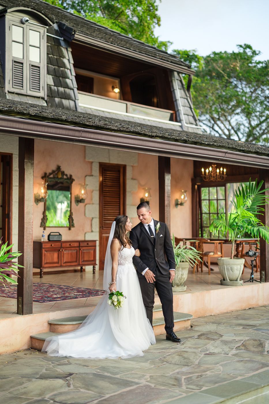 Bride and groom share a romantic moment on a stone patio outside a tropical colonial-style wedding venue.
