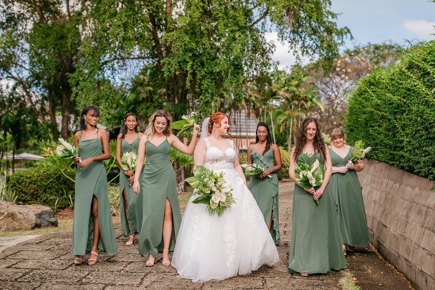 Bride in white gown with bridesmaids in sage green dresses holding bouquets outdoors at a garden wedding.