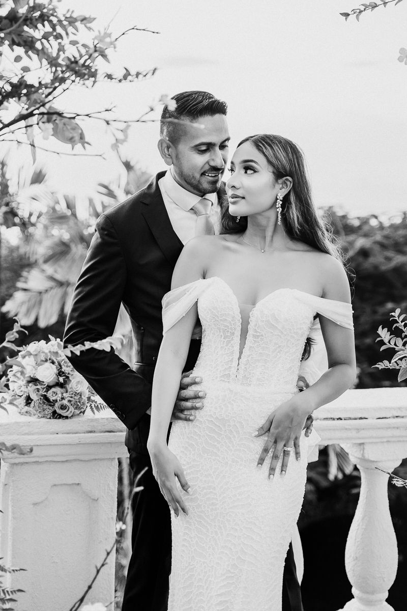Black and white wedding photo of bride in off-shoulder gown and groom in suit embracing outdoors.