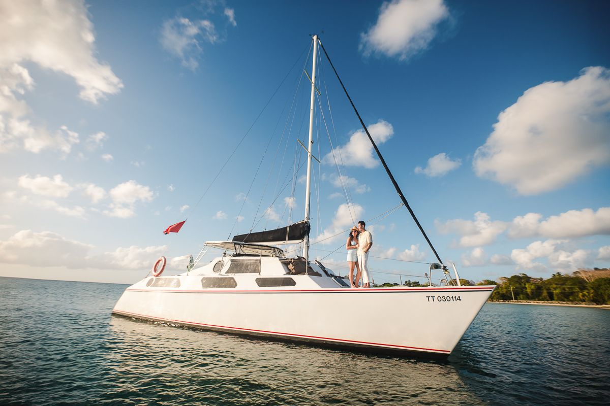Couple standing on a white catamaran sailboat anchored in calm turquoise Caribbean waters under a bright blue sky.