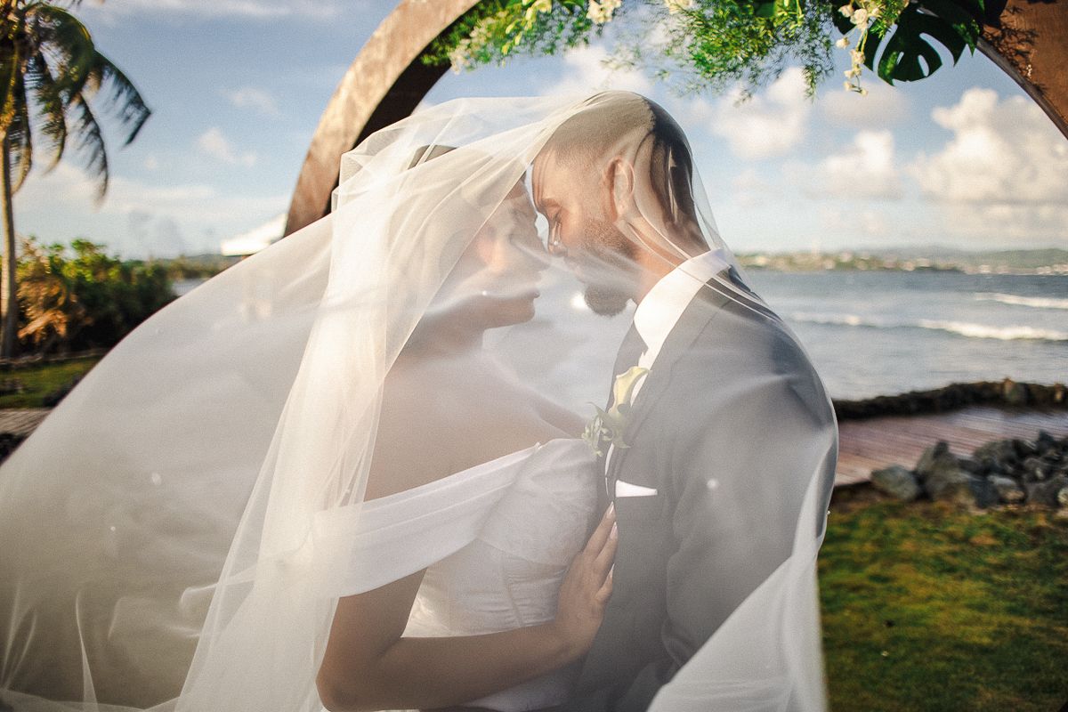 Bride and groom share romantic moment under flowing veil at oceanside wedding ceremony in tropical setting.