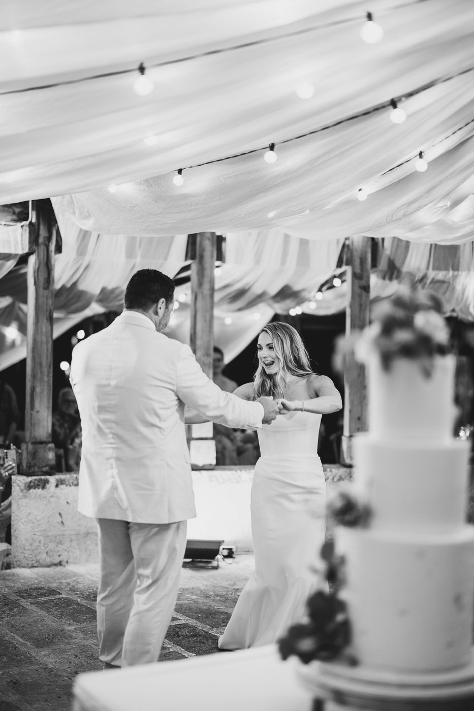 Black and white photo of bride and groom sharing first dance under draped fabric and string lights with wedding cake nearby.