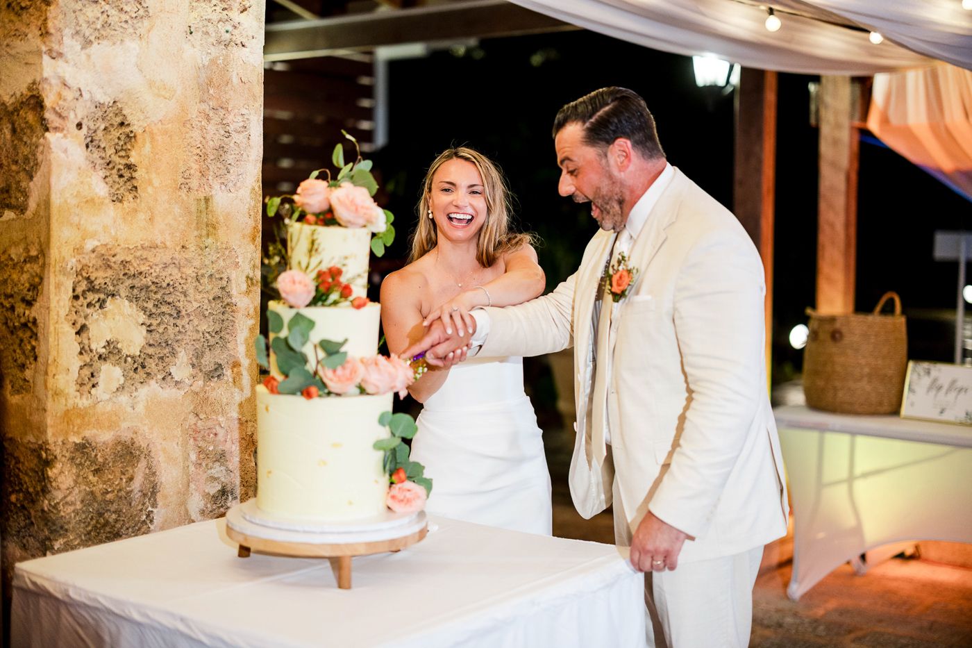 Bride and groom smiling while cutting a white floral wedding cake at an outdoor evening reception.