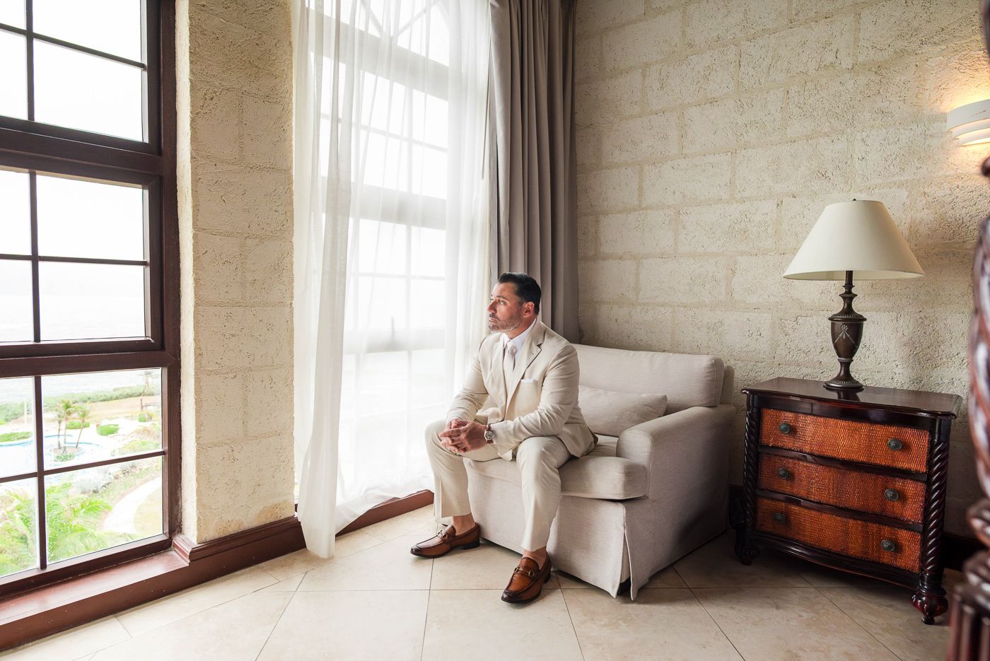 Man in white suit sitting on sofa by large window in elegant room with lamp and wooden dresser.