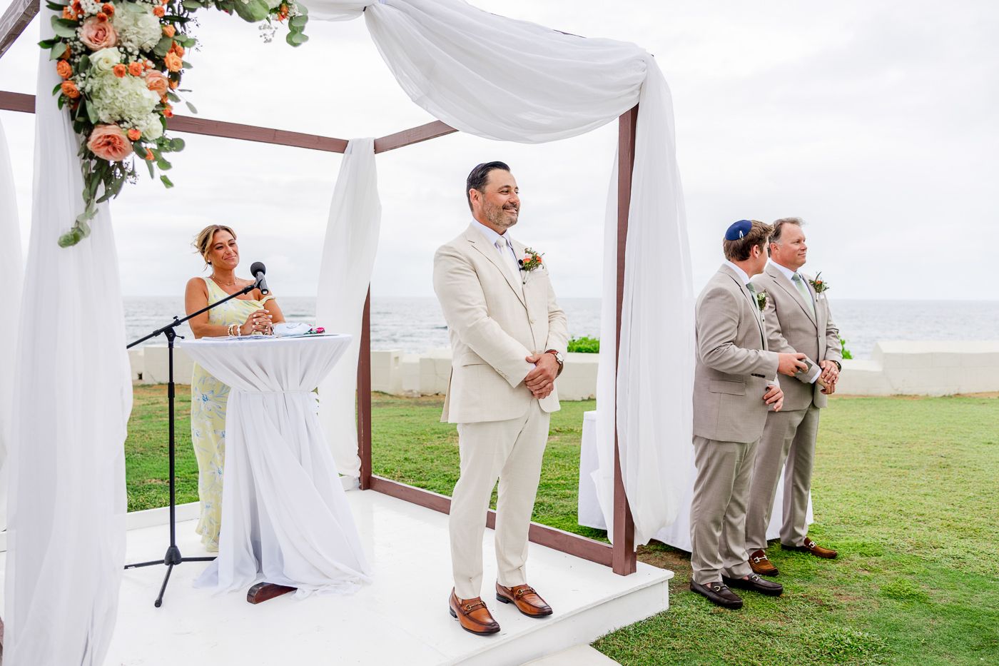 Groom in white suit stands at outdoor beach wedding ceremony under draped floral arch with officiant and groomsmen.