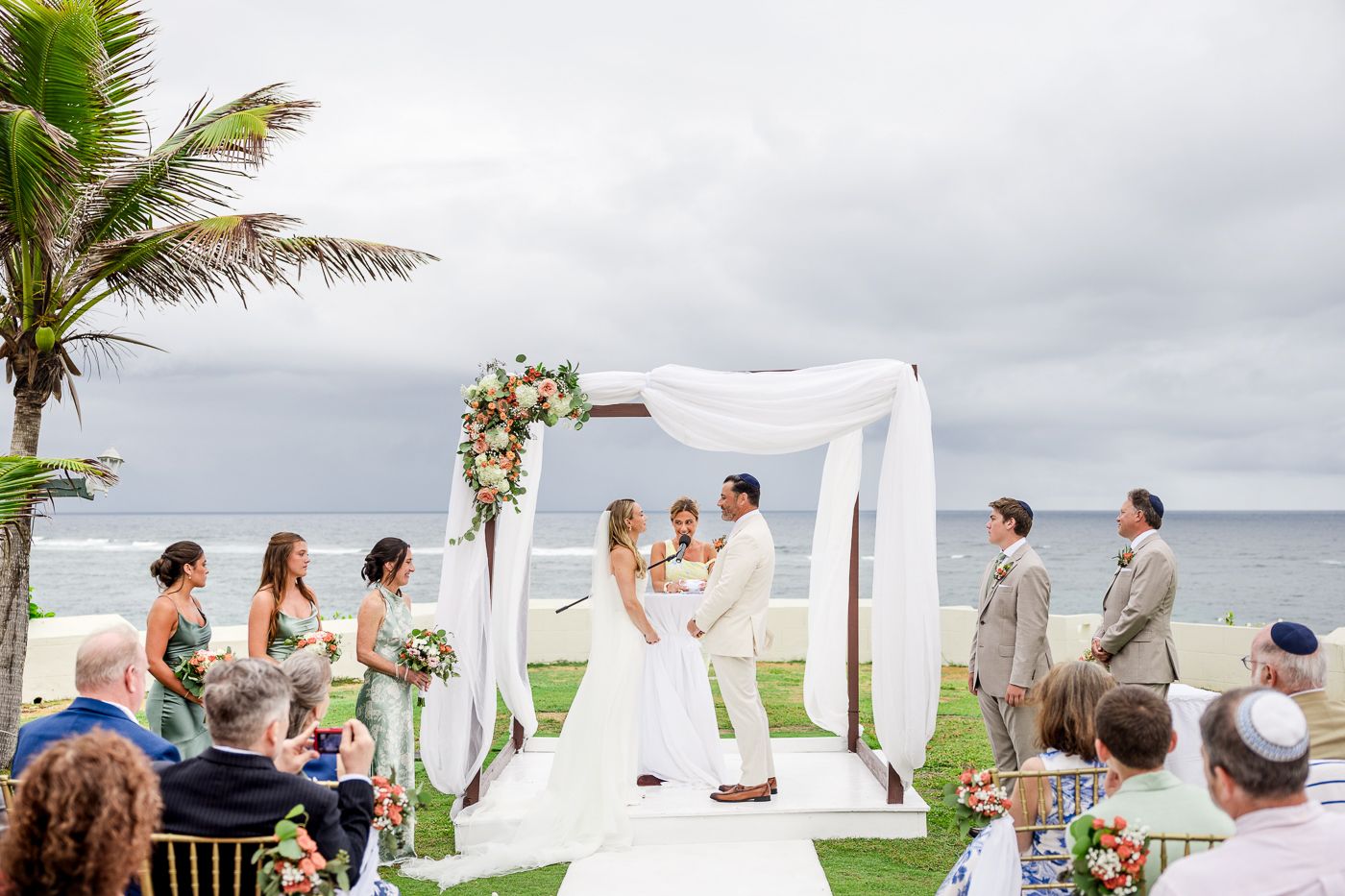 Outdoor beach wedding ceremony under white draped arch with floral decor, bridal party, and ocean backdrop.