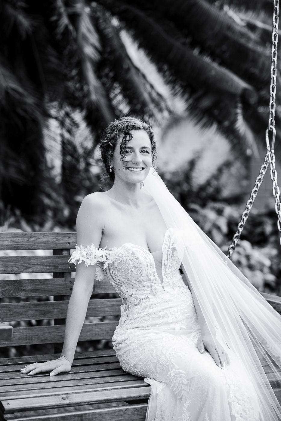 Black and white portrait of bride sitting on wooden bench with flowing veil.