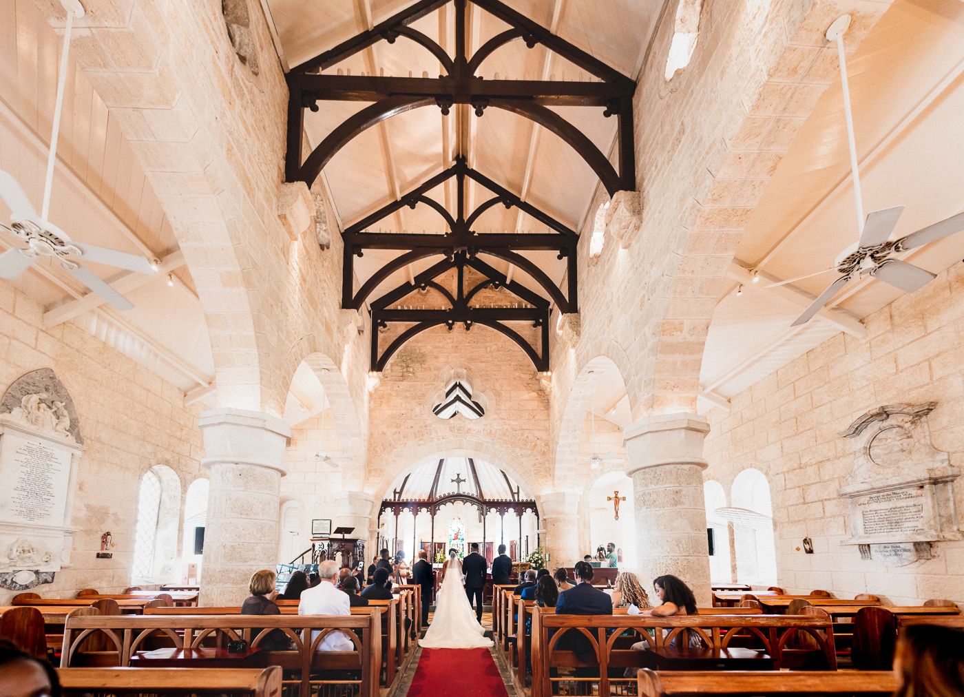 Interior view of a church wedding ceremony with exposed wooden beams and stone columns along a red-carpeted aisle.