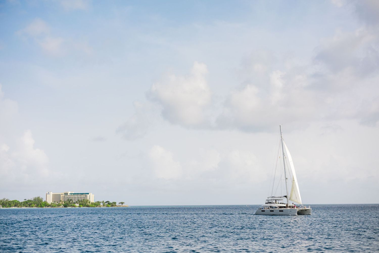 White sailboat glides across barbados blue waters near tropical shoreline under partly cloudy sky. Carlisle Bay, barbados