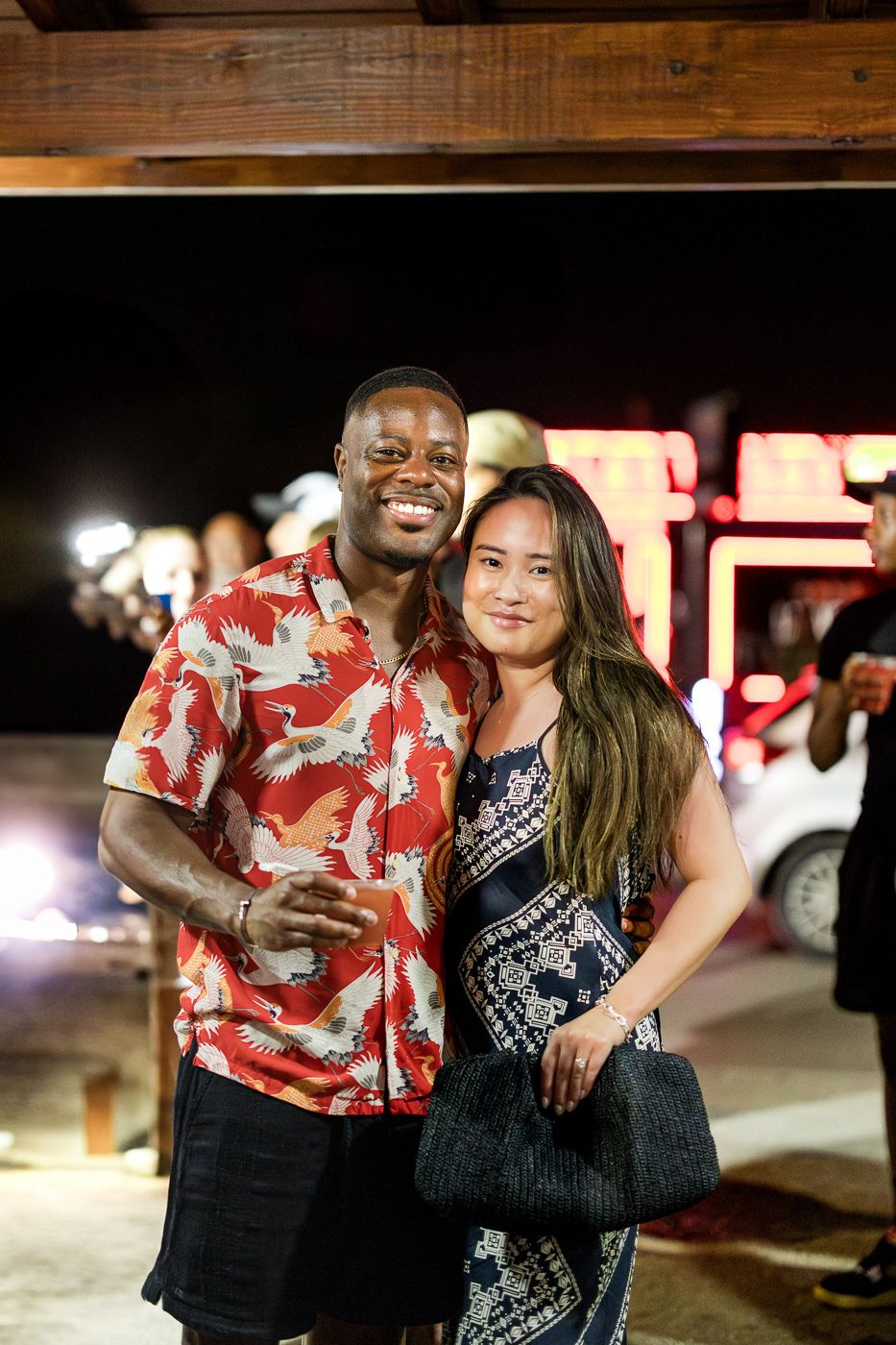 Couple in festive attire smiling at night with illuminated signage in background.