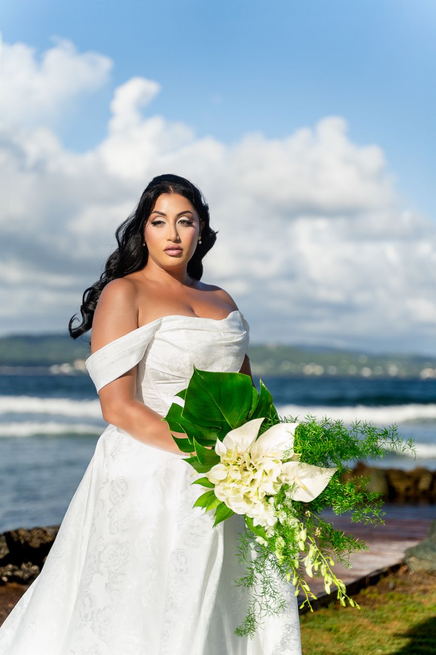 White wedding gown and bouquet captured against scenic ocean backdrop with blue sky and clouds.