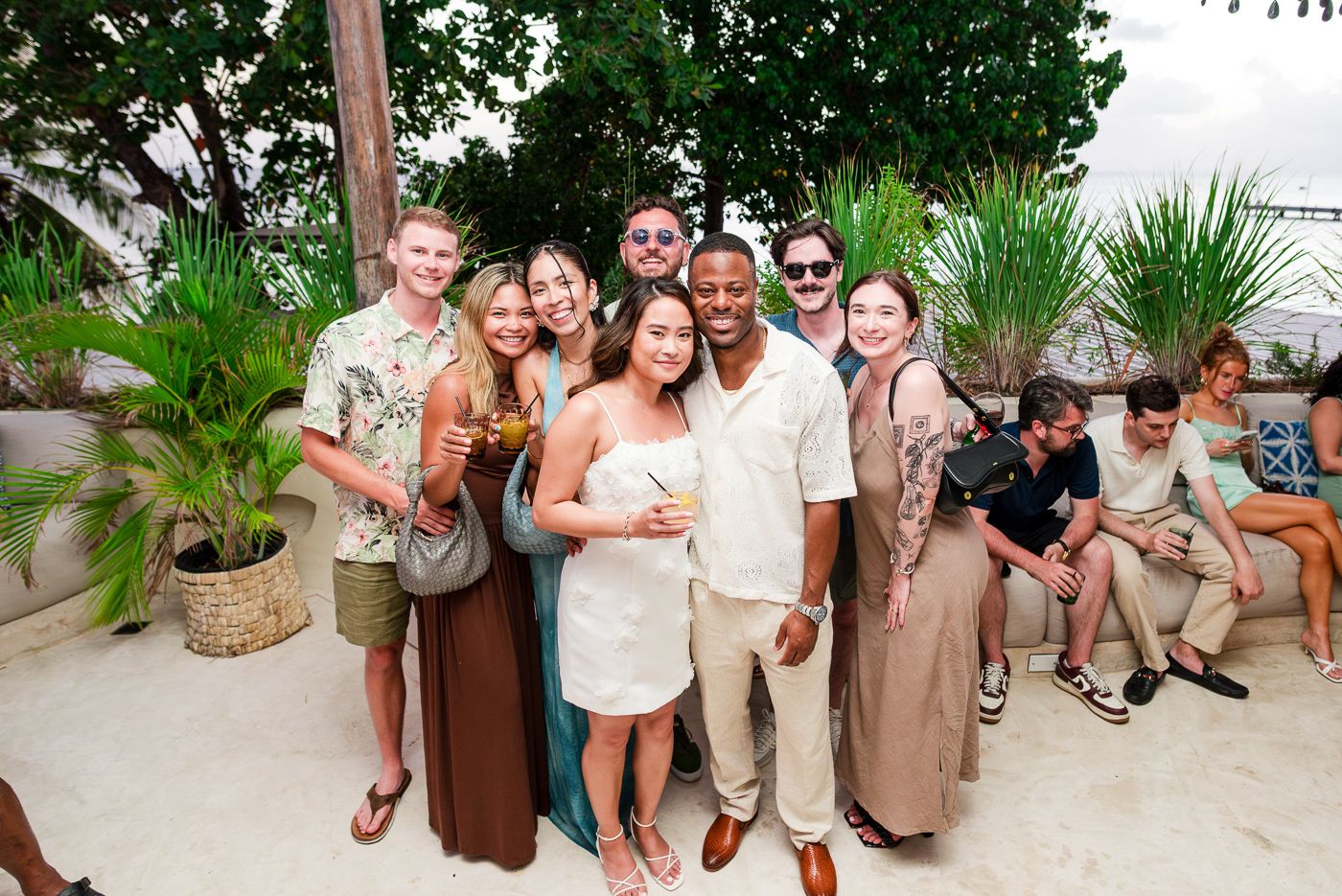A group of wedding guests pose together for a casual photo at an outdoor tropical venue.