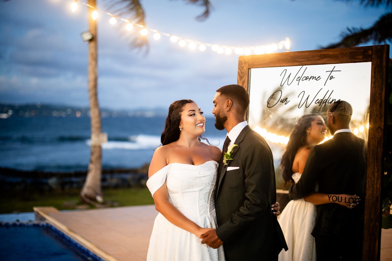 String lights illuminate an evening beach wedding reception under palm trees.