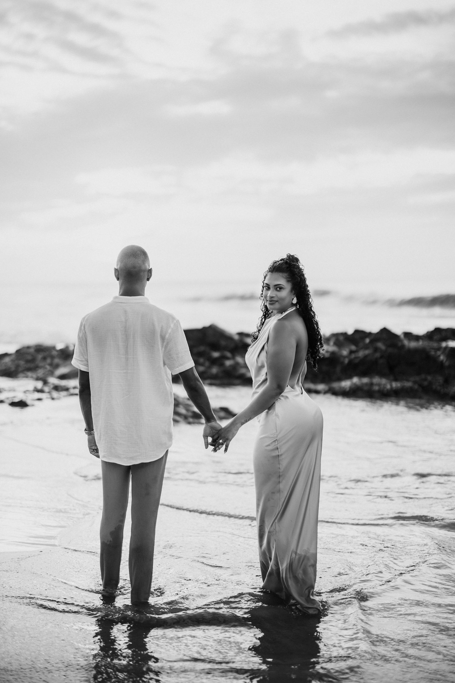 Black and white photo of a couple holding hands at the beach, woman in elegant dress looking back at camera.