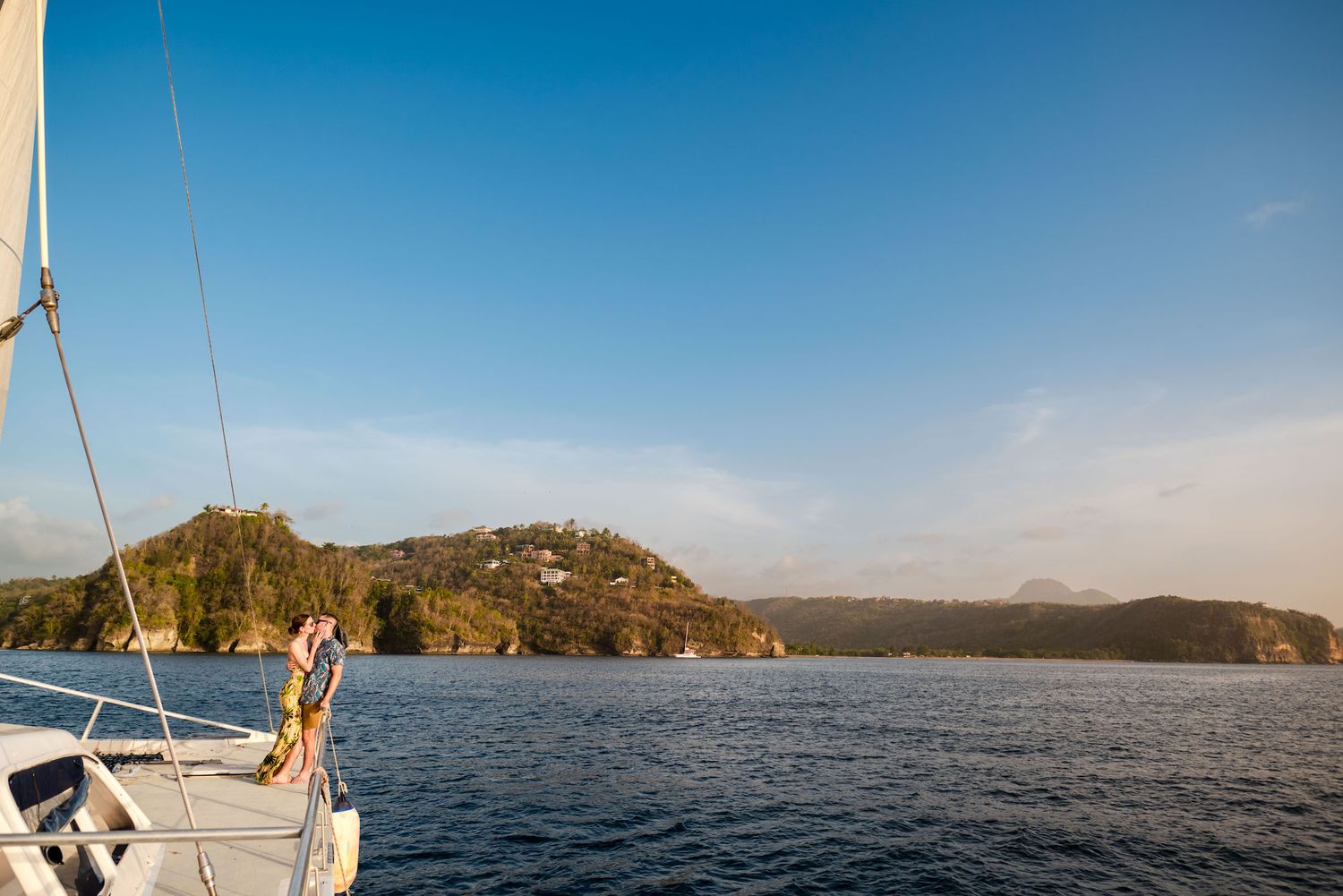 Person standing on the bow of a sailboat with lush green island hills and calm blue waters under a clear sky.