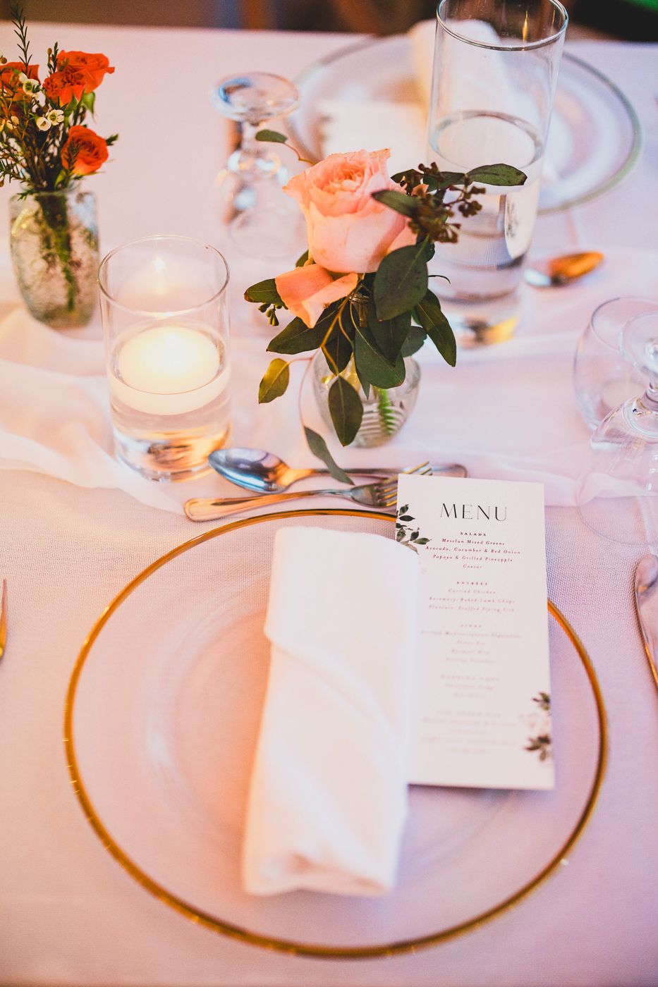 Elegant wedding table setting with gold-rimmed plate, white napkin, menu card, peach roses, and candle centerpiece.