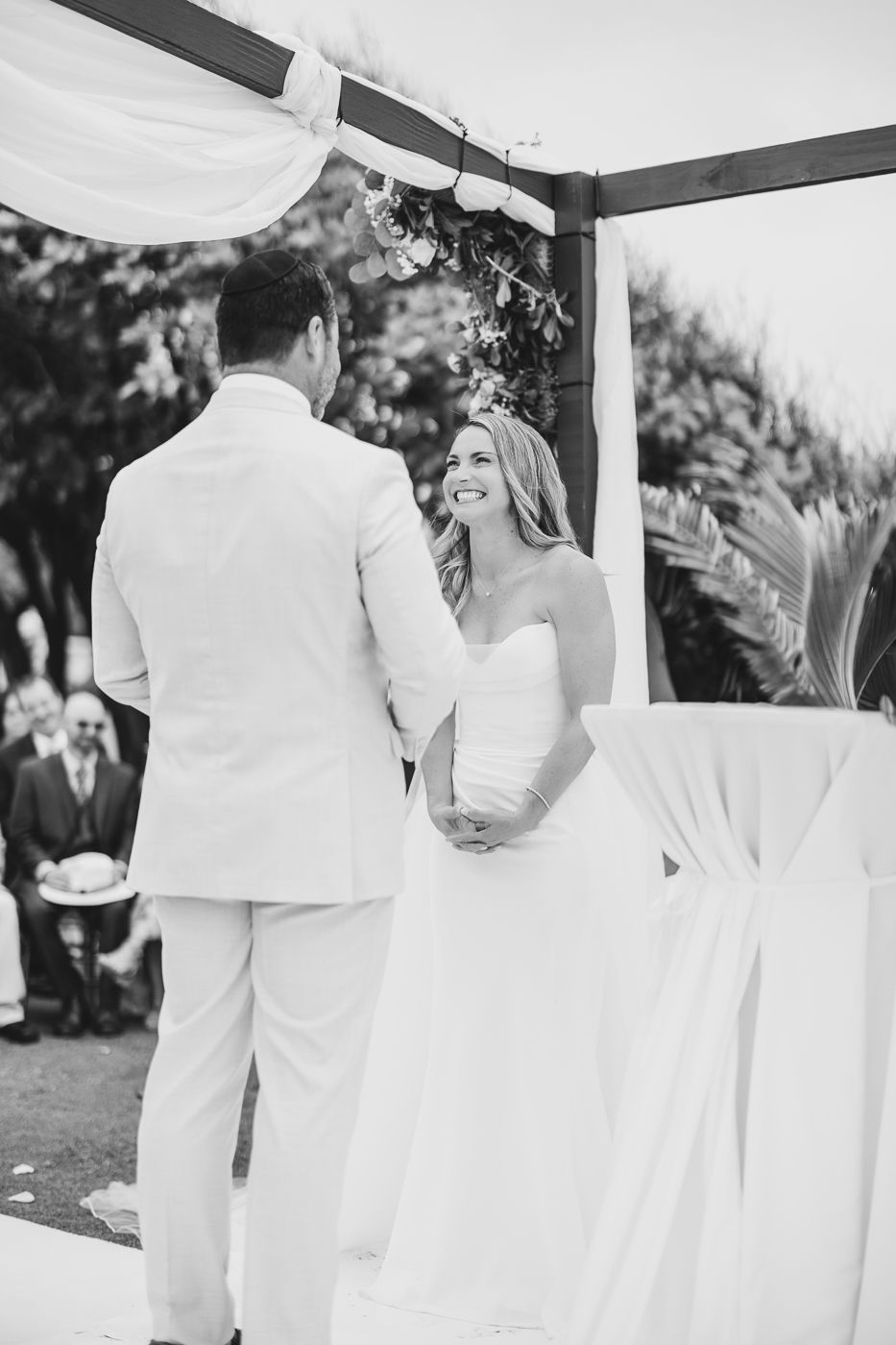 Black and white photo of a smiling bride in a strapless gown holding hands with groom at outdoor wedding ceremony.