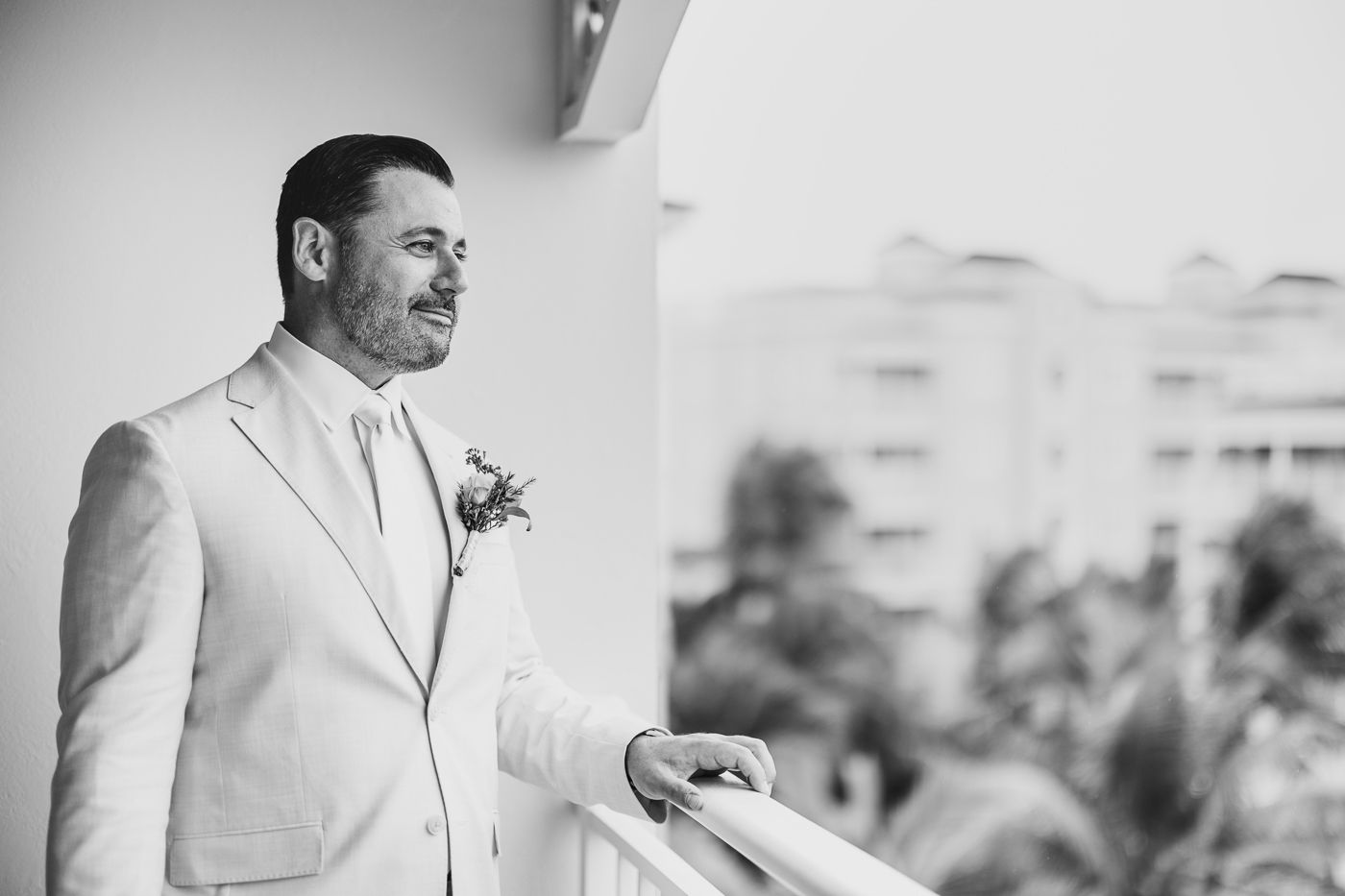 Groom in light suit with boutonniere standing on balcony overlooking tropical scenery.