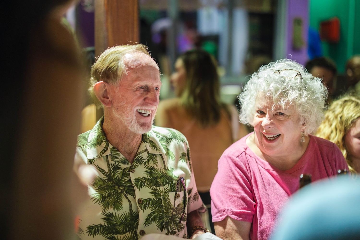 Two elderly people laughing joyfully together at a colorful indoor gathering, man in floral shirt and woman in pink top.