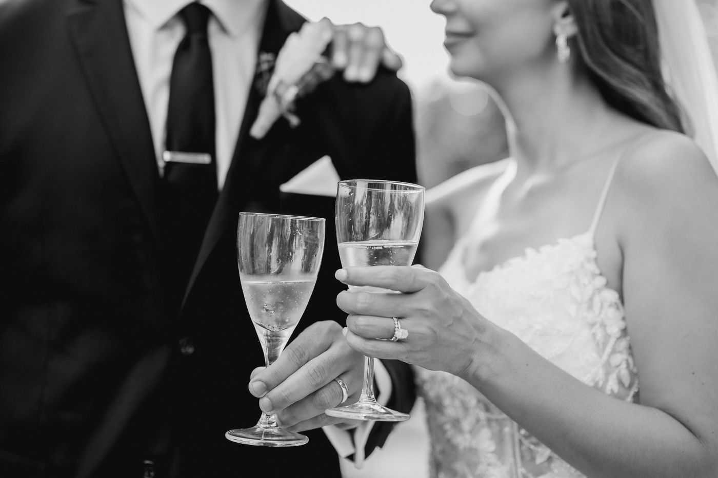 Black and white photo of bride and groom toasting with champagne glasses, bride wearing engagement ring and lace dress.