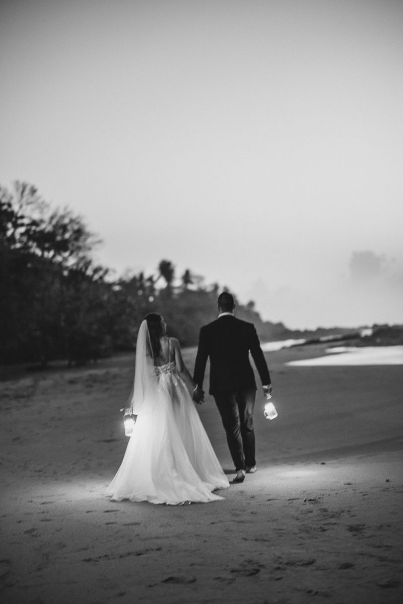 Black and white photo of bride and groom walking on beach holding lanterns at dusk.
