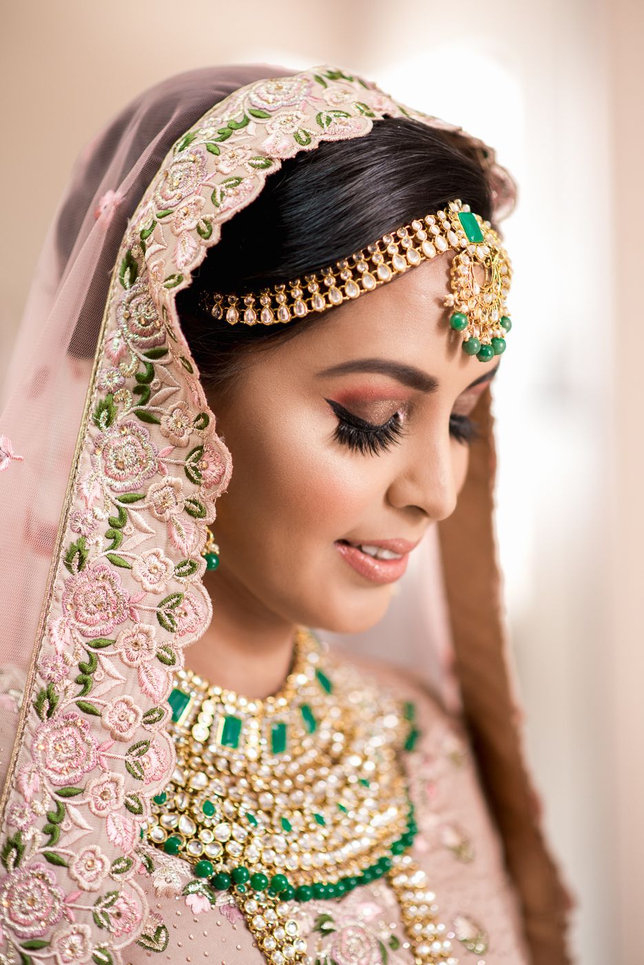 Indian bride in pink lehenga with green and gold jewelry, maang tikka, and embroidered dupatta, looking down gracefully.