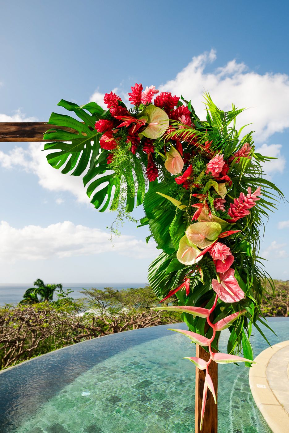 Wedding arch decorated with red tropical flowers and monstera leaves overlooking ocean vista.