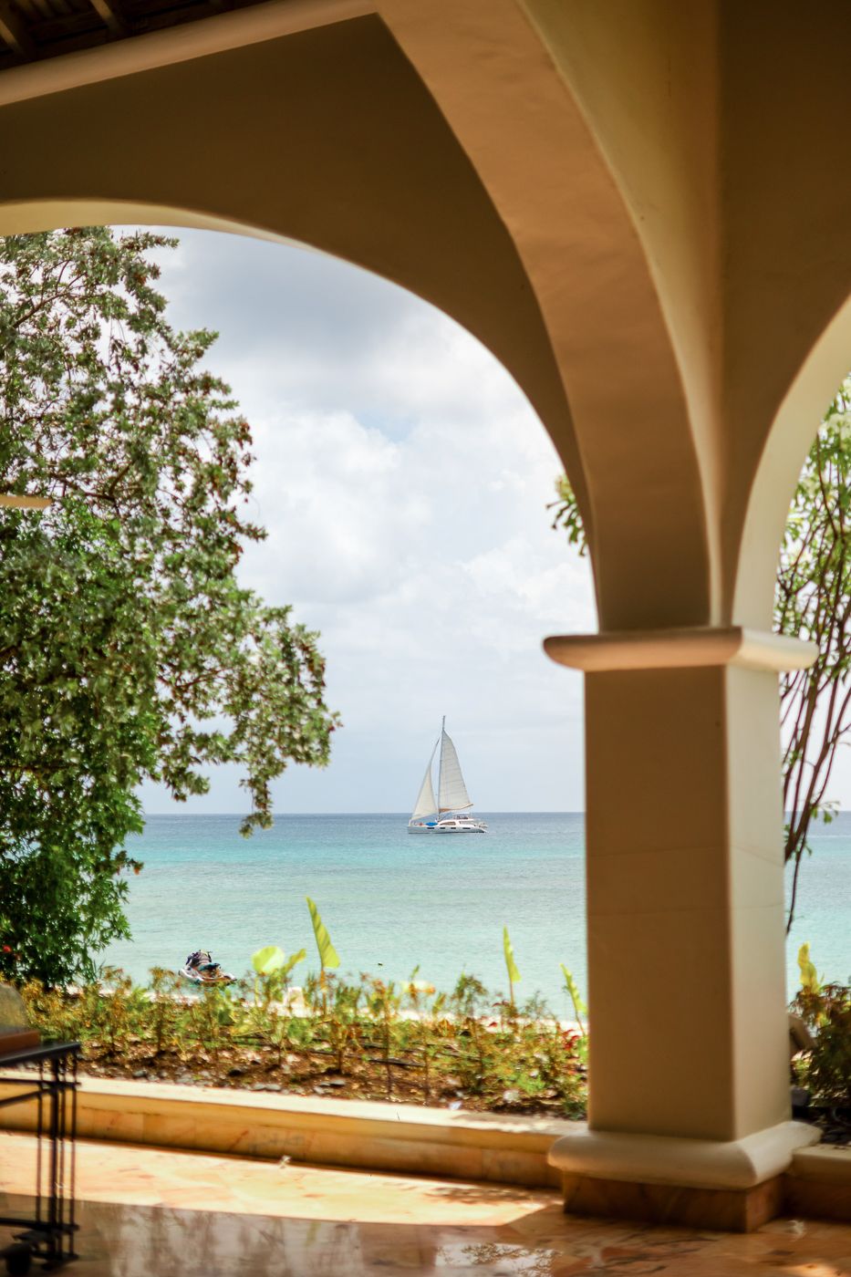 View of a sailboat on turquoise waters through an elegant archway with tropical plants framing the scene.