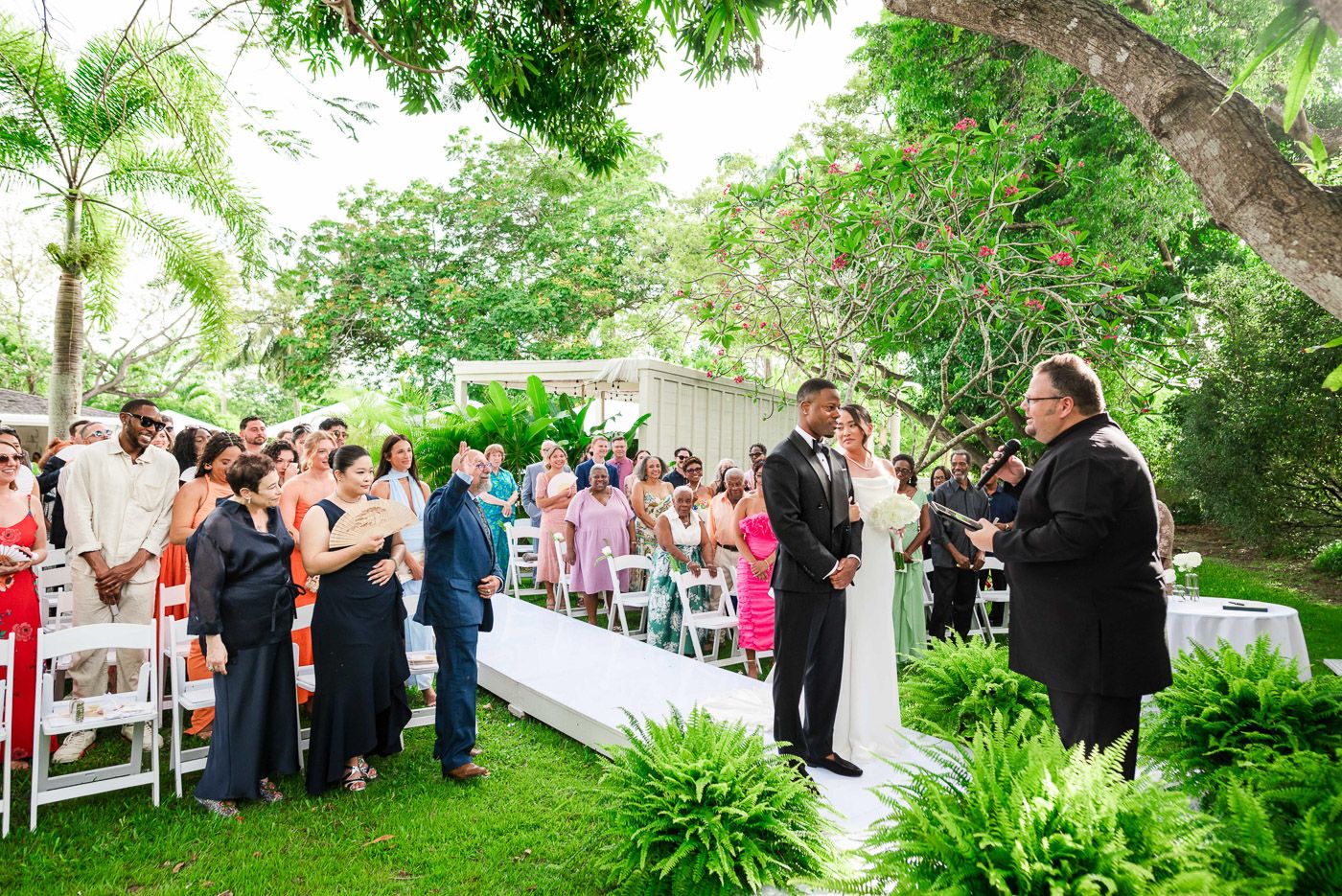 An outdoor wedding ceremony surrounded by trees and white chairs with guests watching the proceedings.