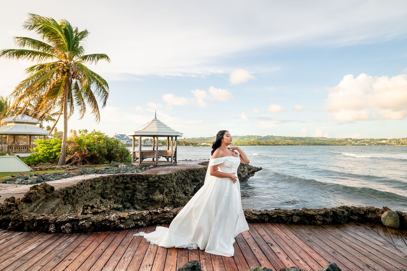 Bride in flowing white dress poses along tropical beachfront with palm trees and ocean views.