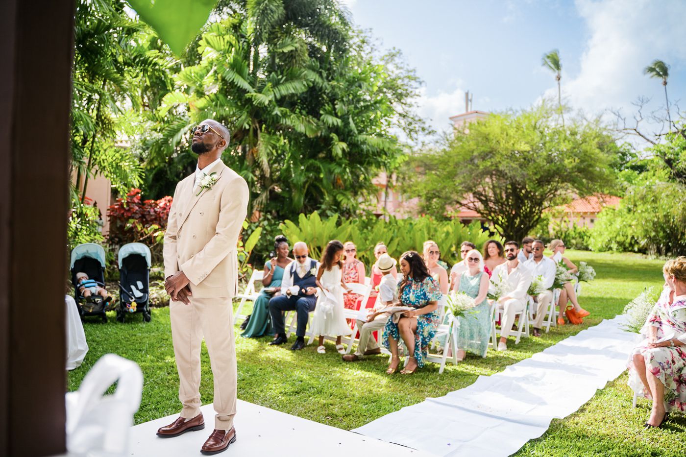 An outdoor wedding ceremony setup with white chairs on a lawn surrounded by tropical landscaping.