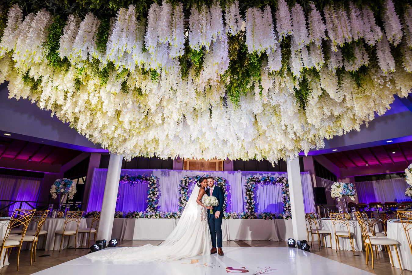 Bride and groom share first dance under stunning white wisteria floral ceiling installation in elegant wedding venue.