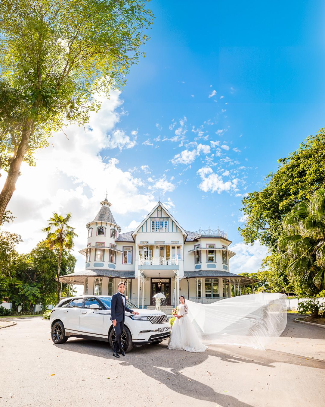 Bride and groom pose by white Range Rover before grand Victorian mansion under blue sky.