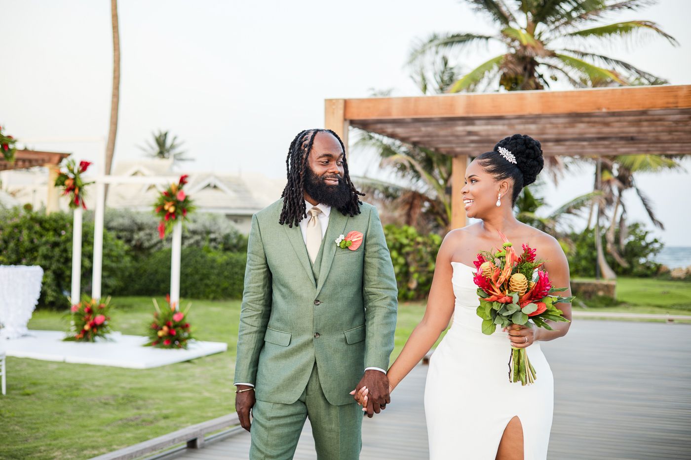 Bride and groom holding hands in tropical wedding setting, groom in sage suit, bride in white dress holding vibrant bouquet.