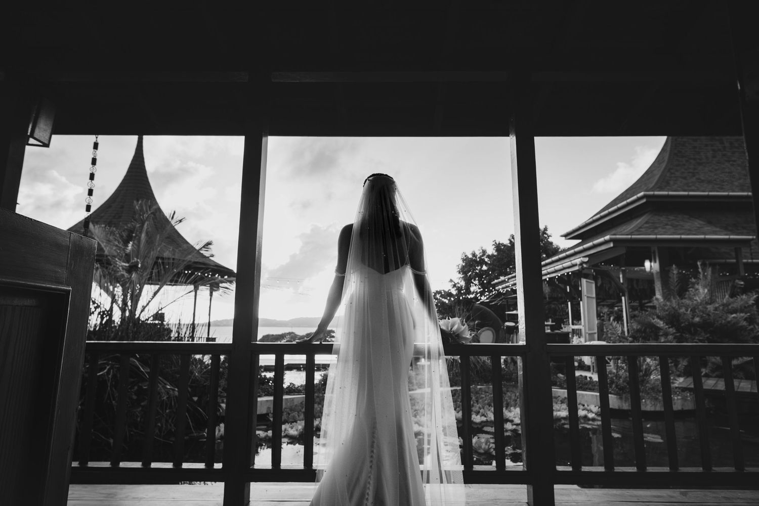 Black and white photo of a bride in a flowing gown and long veil standing on a balcony overlooking traditional pavilion rooftops.