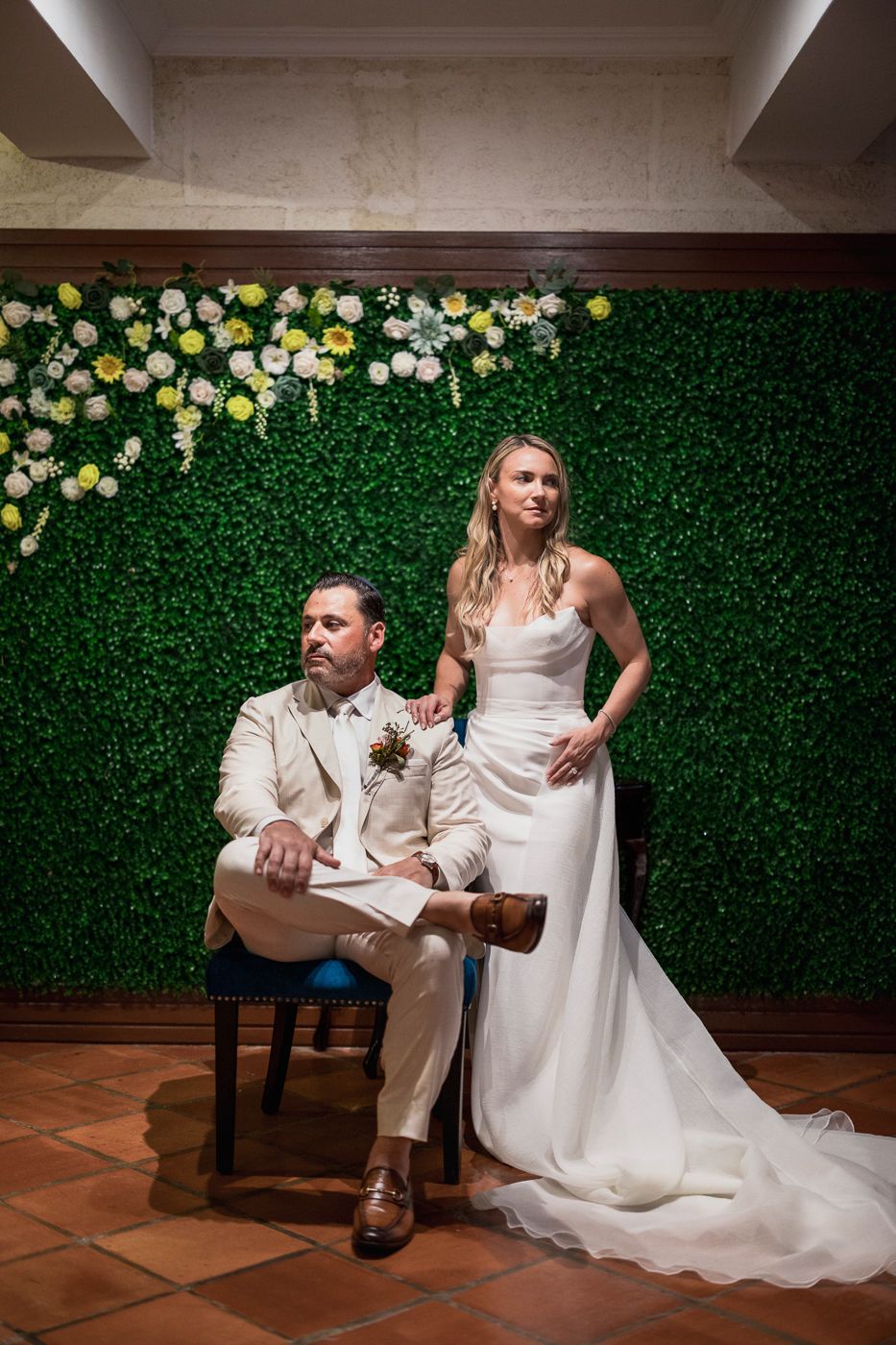 Bride and groom pose elegantly before a lush green floral wall adorned with yellow and white flowers.