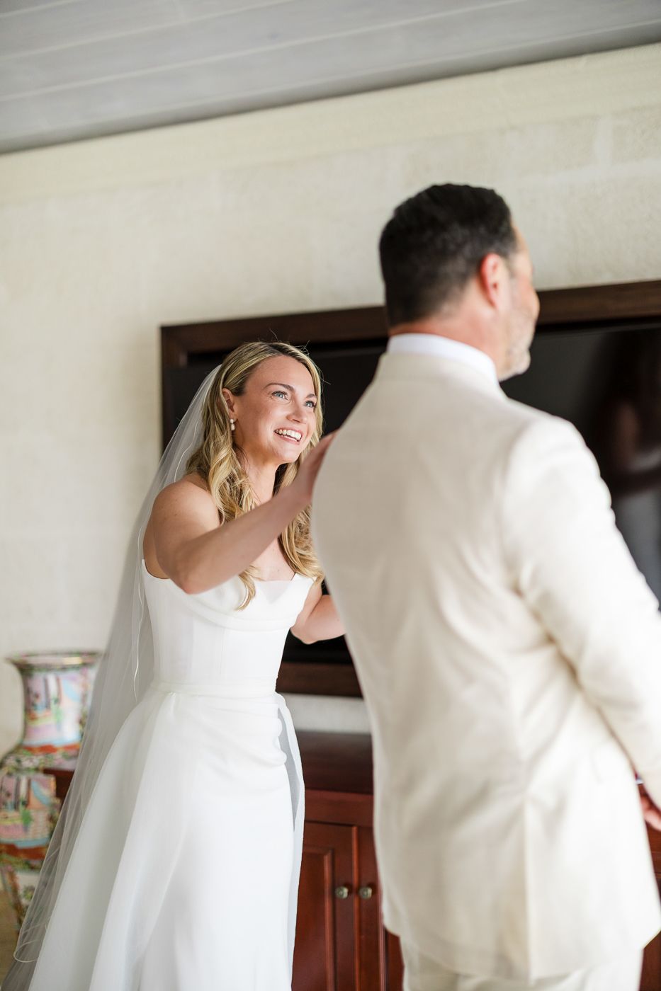 Bride in white gown and veil smiles at groom in cream suit during an emotional first look moment indoors.