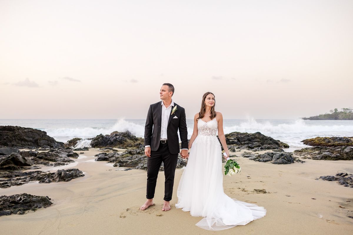 Bride and groom holding hands on rocky beach with ocean waves at sunset.