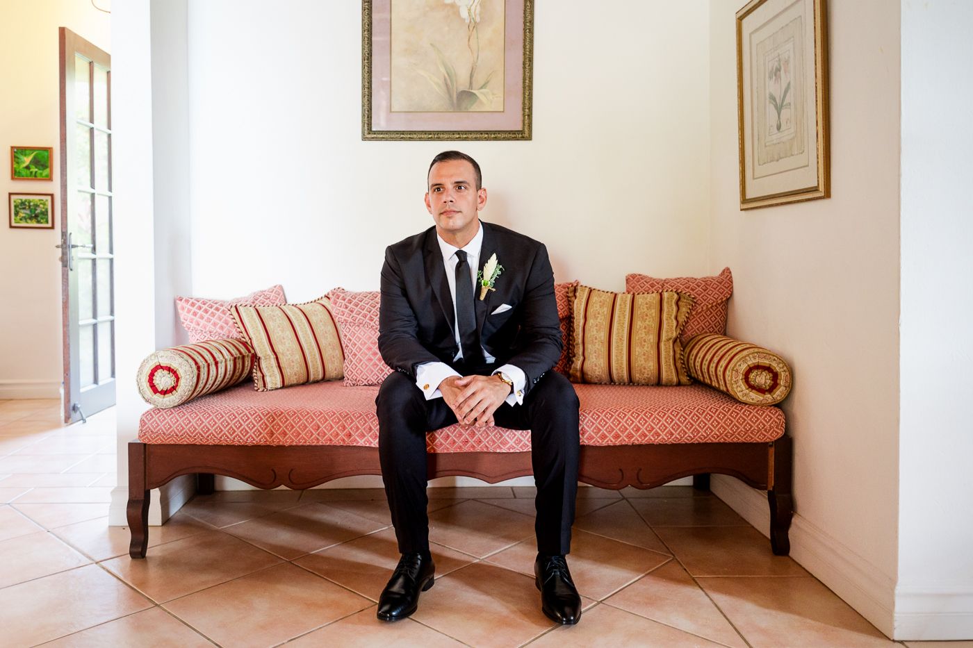 Groom in black suit sitting on red sofa with striped pillows, wearing boutonniere, in elegant room.