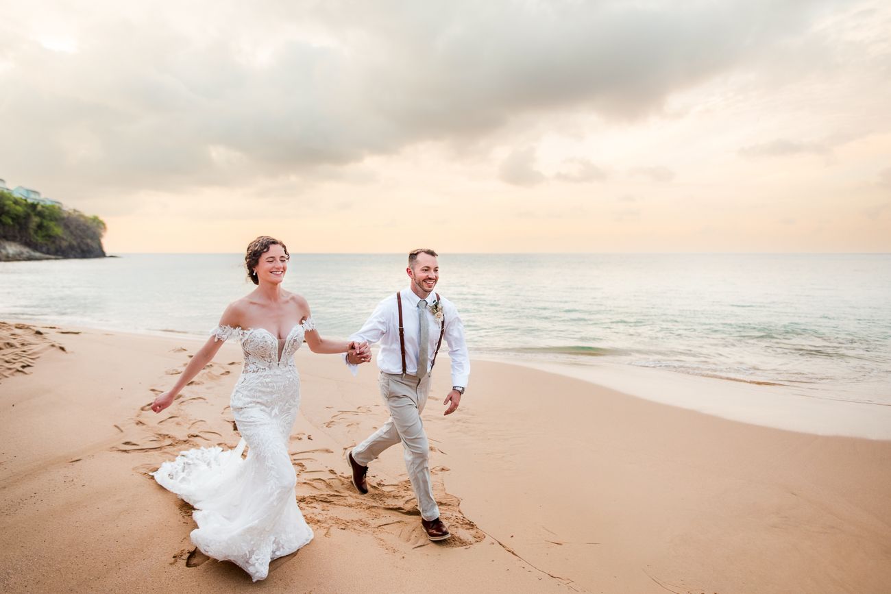 Newlywed couple holding hands and walking joyfully on a sandy beach at sunset in wedding attire.