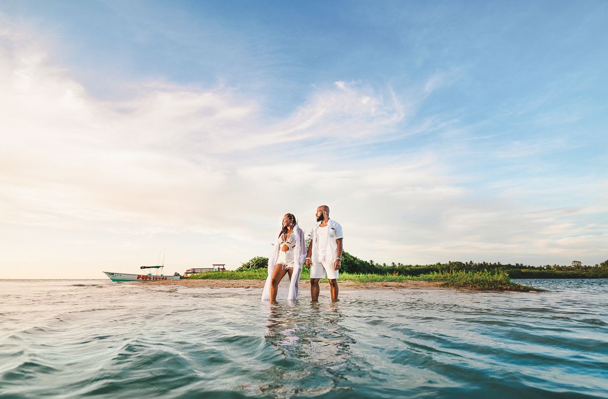 Couple in white outfits standing in shallow tropical ocean water near a lush green island with a boat in the background.