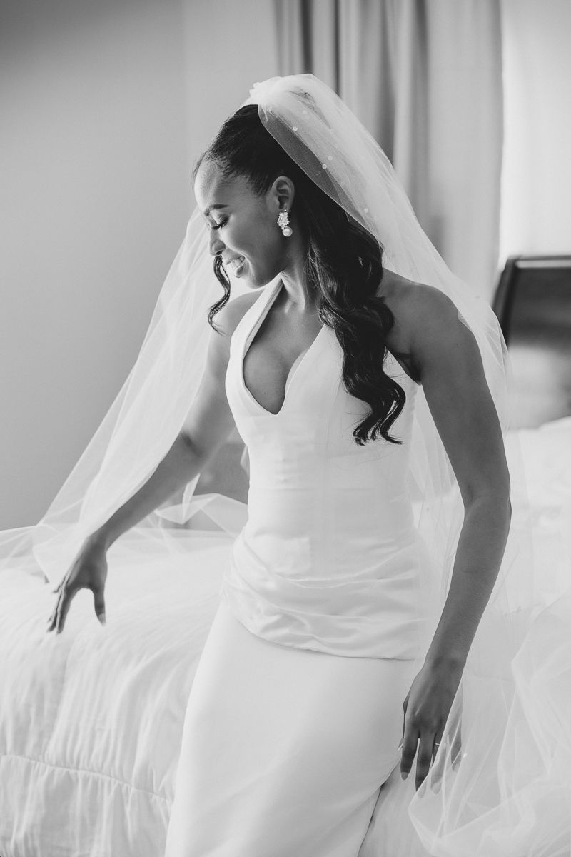 Black and white photo of a smiling bride in a halter wedding gown and veil, sitting on a bed.