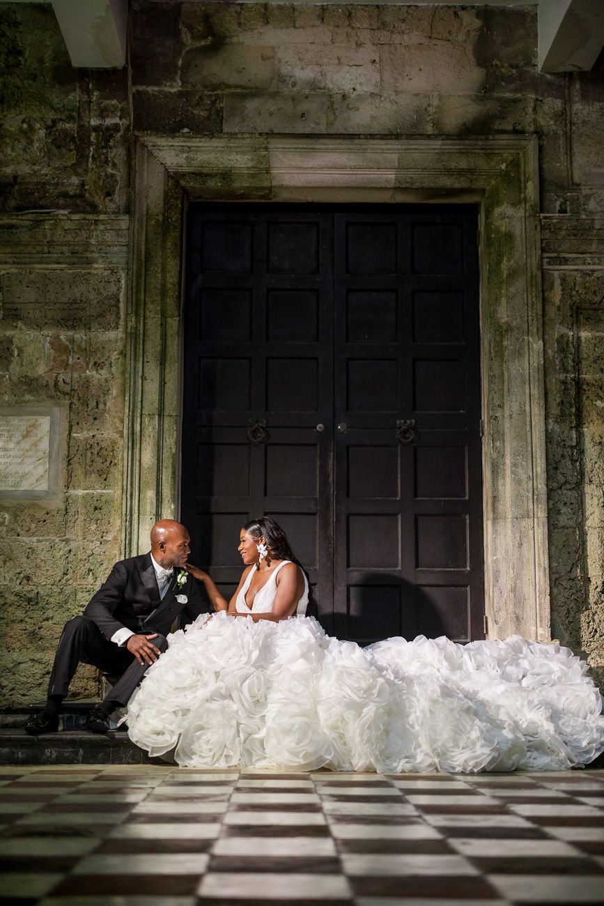 Bride in voluminous white ruffled gown and groom in black tuxedo sit together on checkered floor before ornate dark doors.