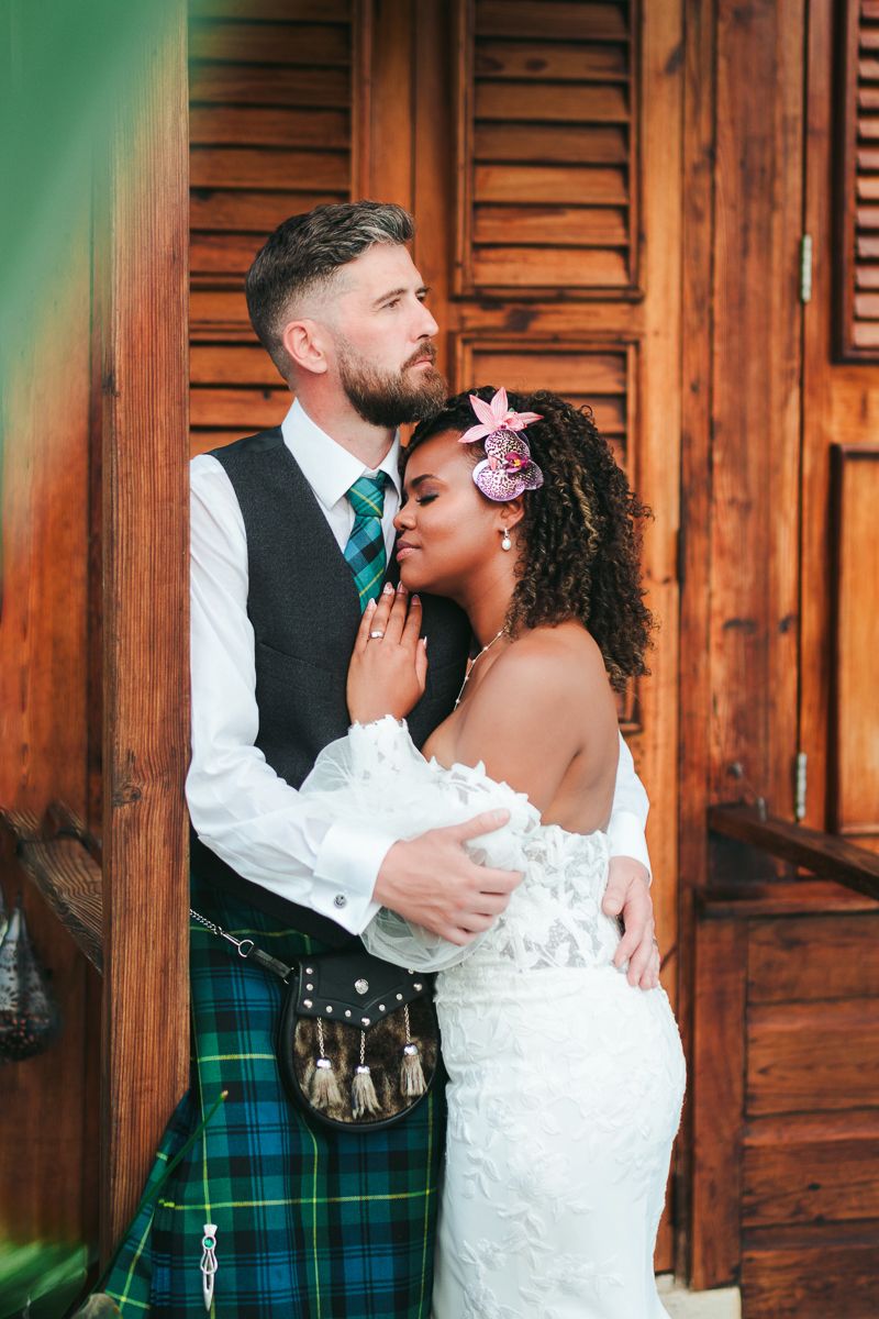 A romantic wedding couple shares intimate moments in front of wooden doors wearing a white dress and green plaid kilt.