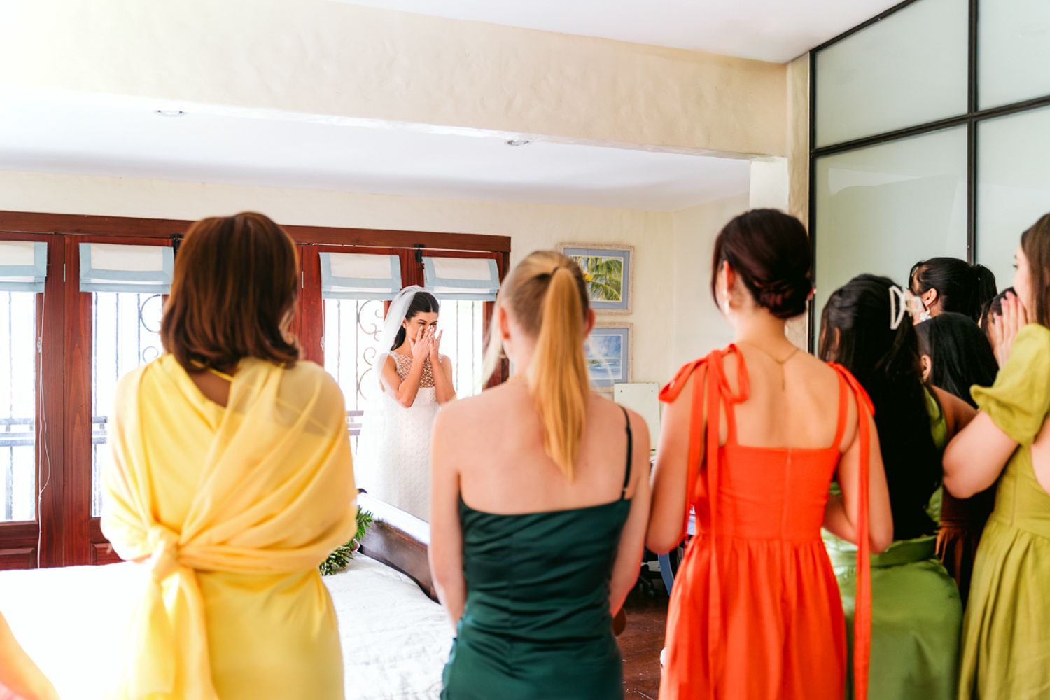 Group of bridesmaids wearing colorful dresses in yellow, green and orange stand together during wedding preparations.