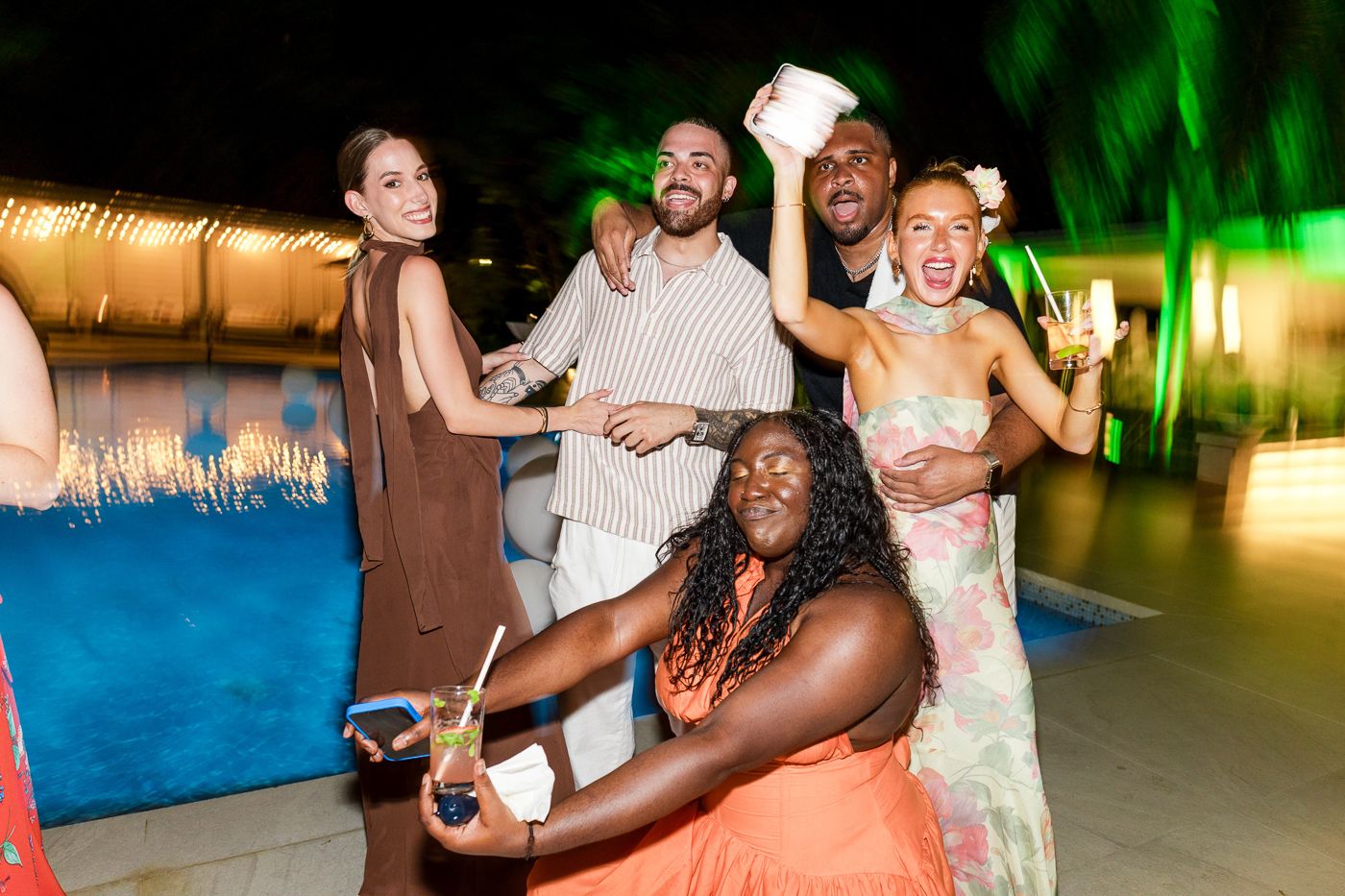 Party guests dancing and celebrating at a poolside evening event with green lighting effects.