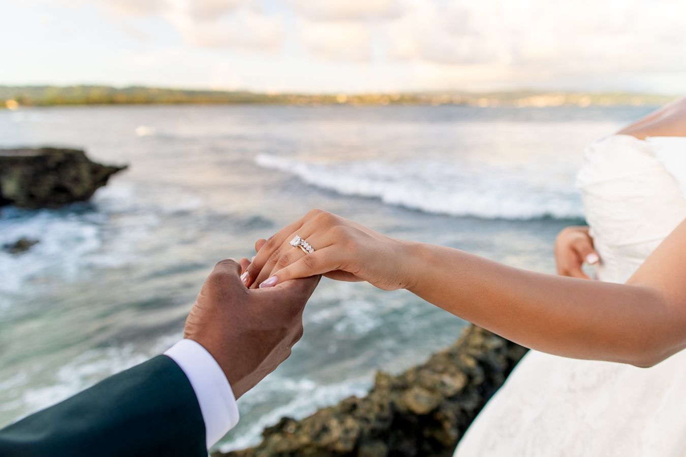 Close up of bride and groom holding hands with ocean waves in background.