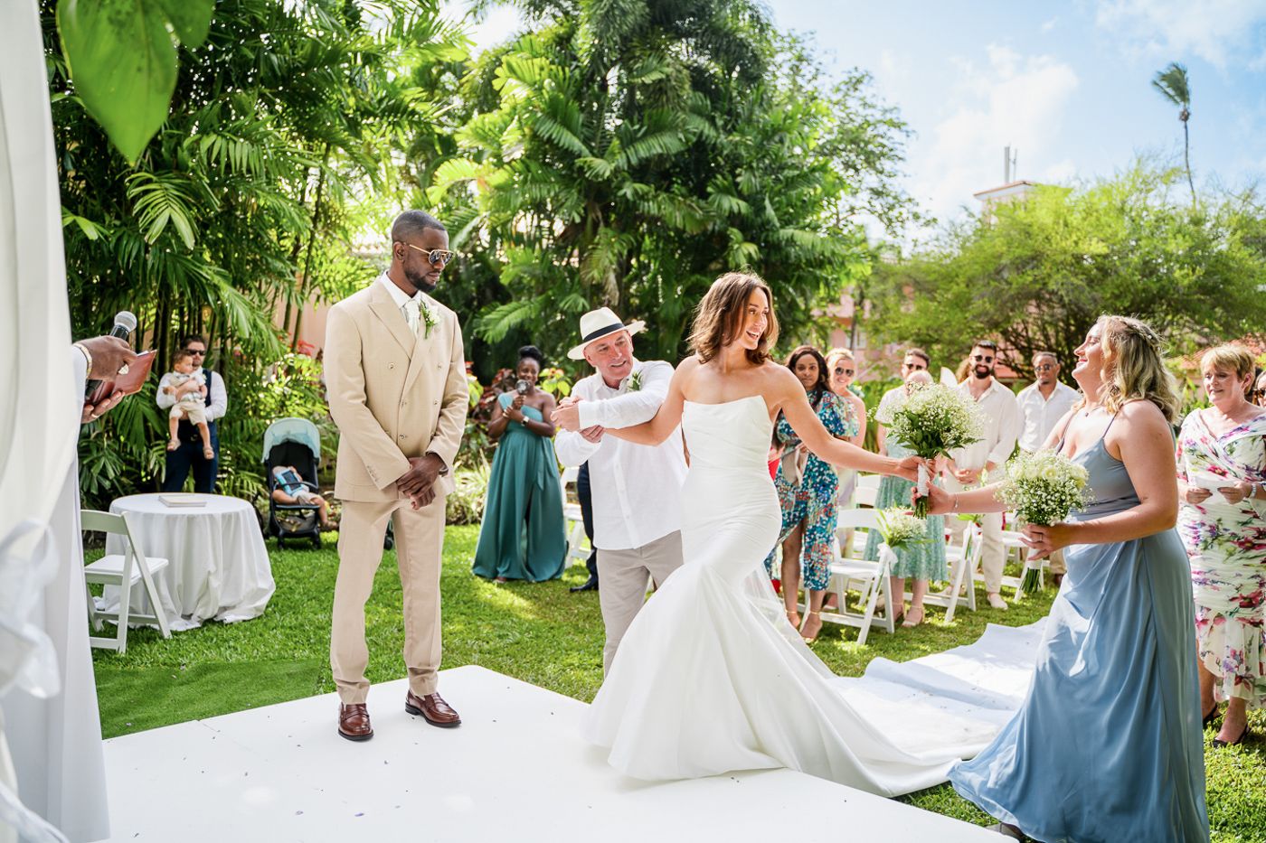 A wedding ceremony in progress on a white aisle runner surrounded by tropical gardens and palm trees.