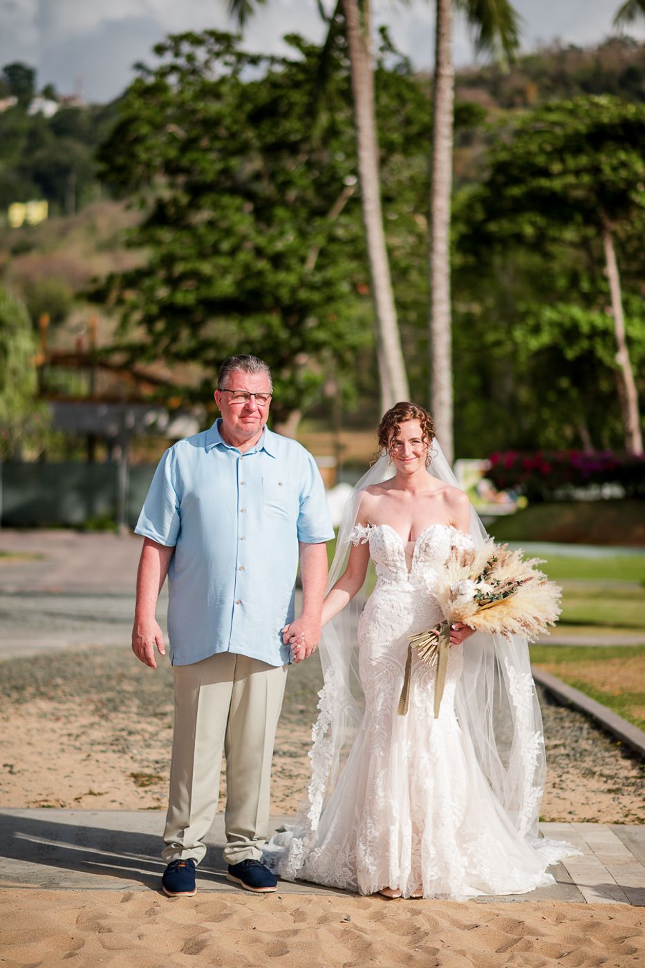 A bride in a white lace dress walks along a beach path with someone in a light blue shirt and khaki pants.