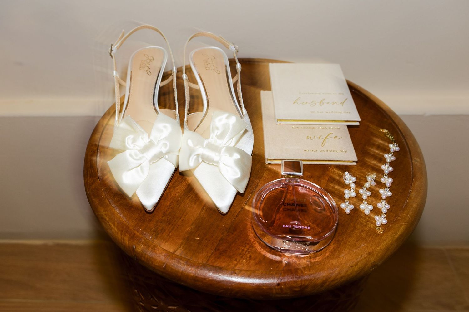 White satin bow bridal heels displayed on wooden tray with perfume bottle, pearl bracelet, and wedding stationery.